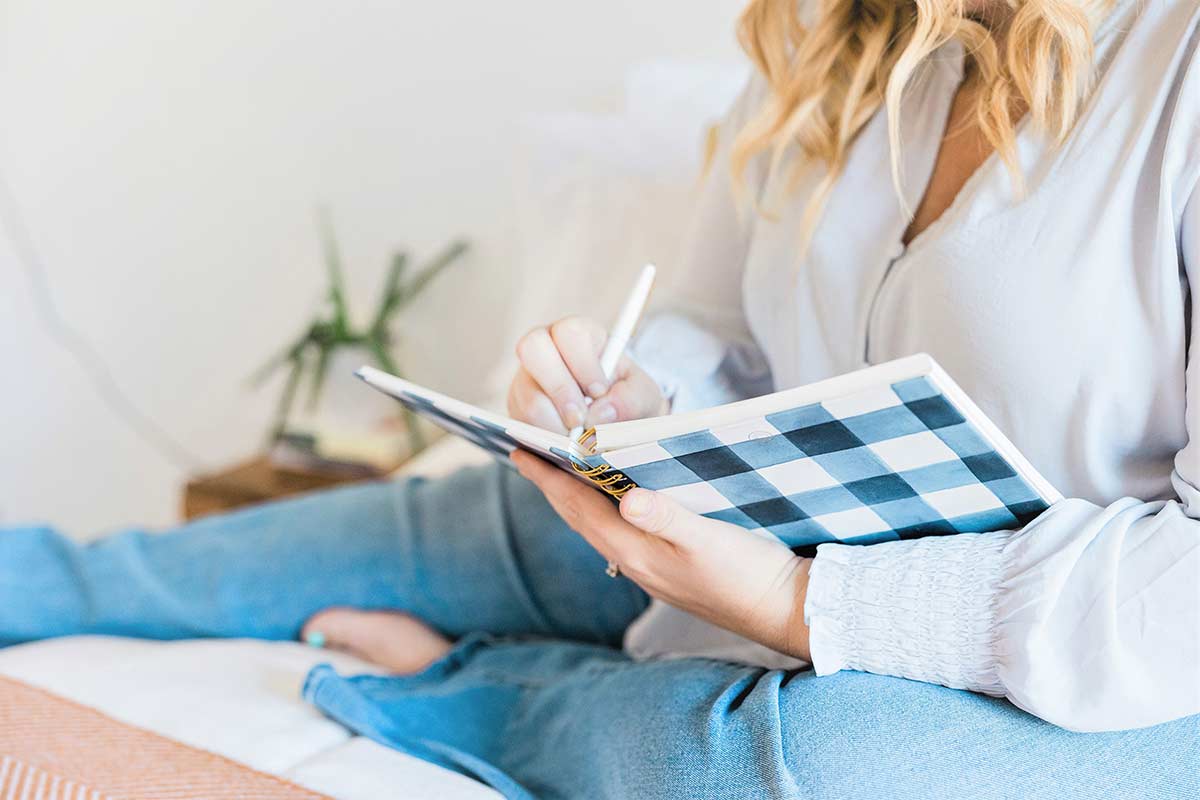 Woman with long blond hair wearing blue blouse and denim jeans sitting and writing about creative journal topics in black and white buffalo check journal.