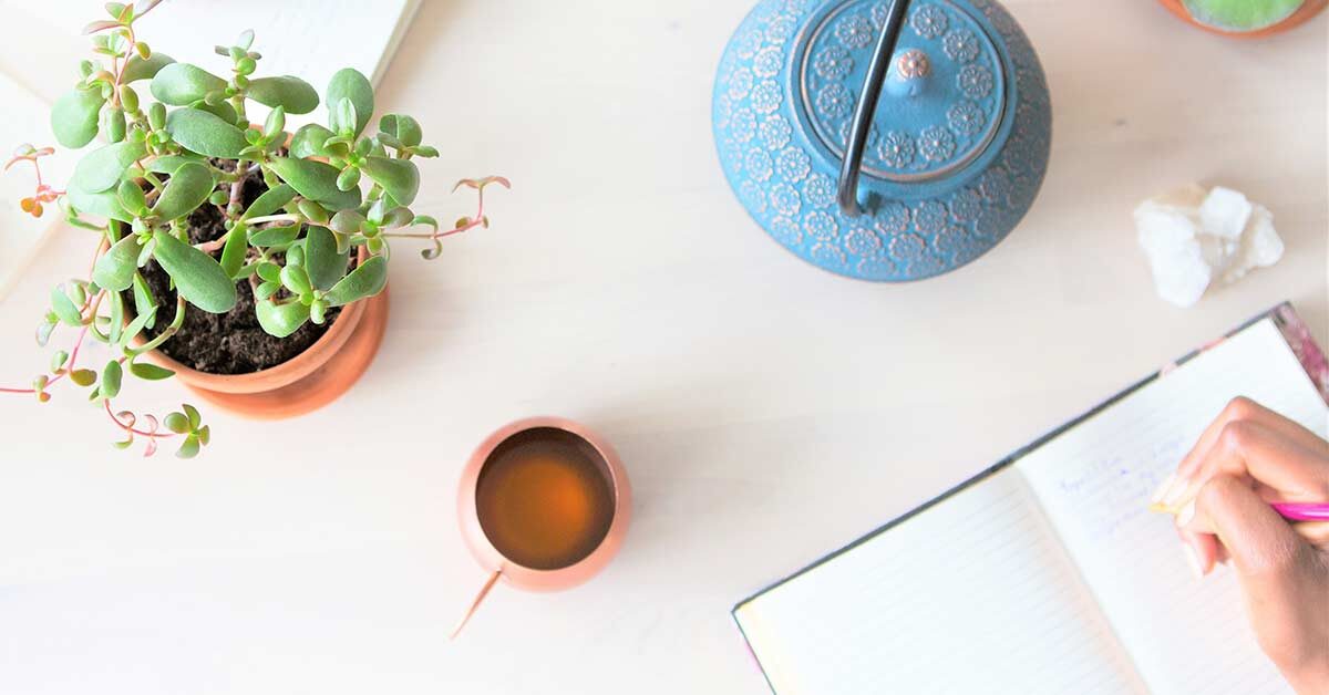 Woman doing creative journaling at white table next to teapot, teacup, and succulent plant.