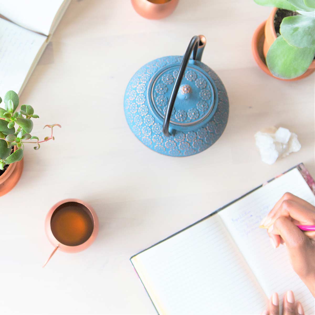 Woman doing creative journaling at white table next to teapot, teacup, and plants.