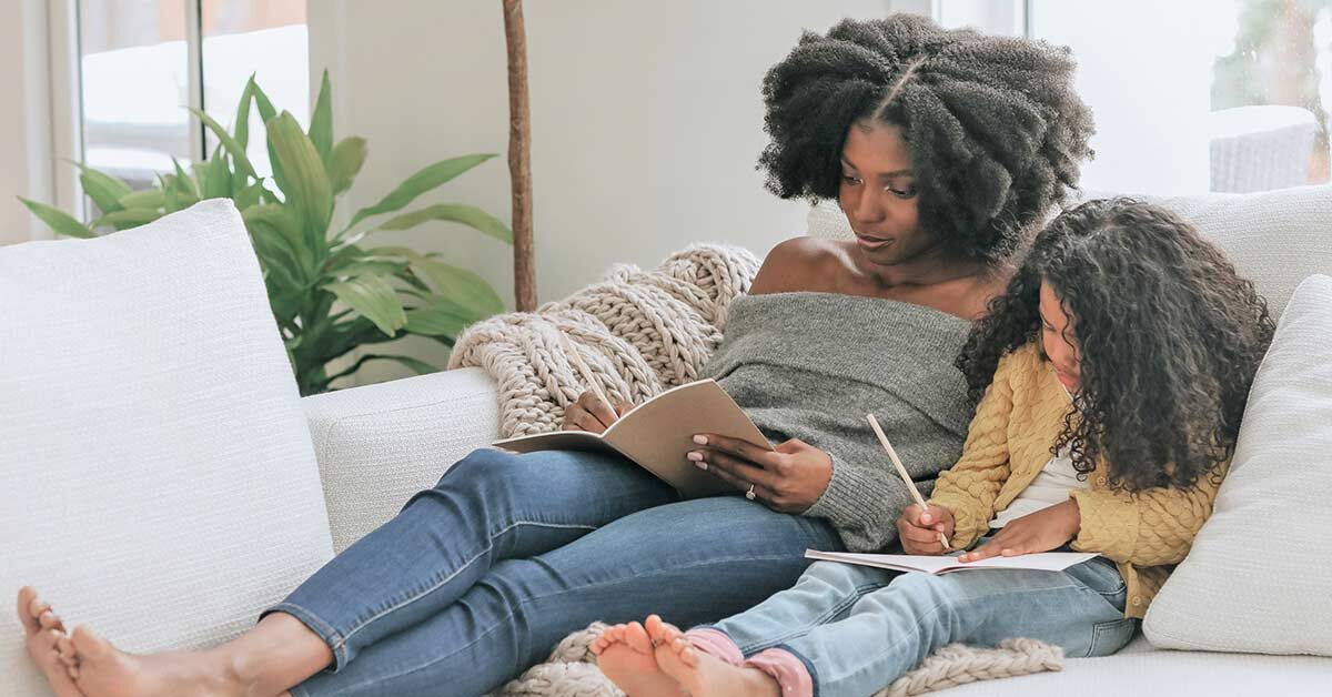 Mother and daughter lounging on white couch and writing in journals.