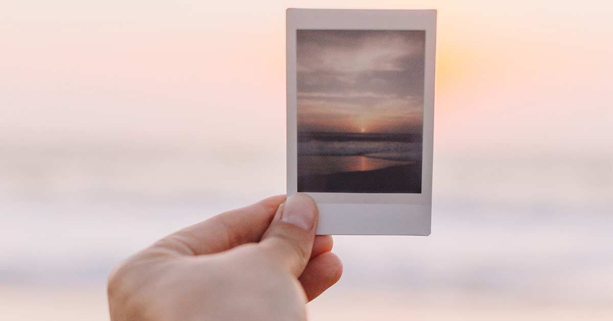 Hand holding up picture of sunset to compare with real sunset.