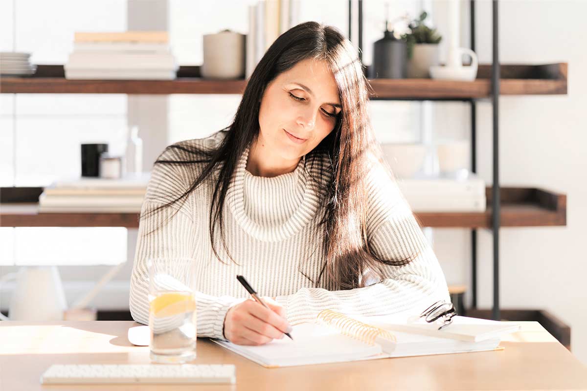 Woman in white sweater with long dark hair doing morning creative writing while sitting at table in sunny room.