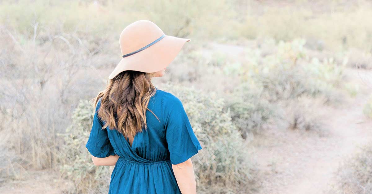 Woman with long hair wearing a floppy hat and blue dress standing in the desert on a slow down Sunday.