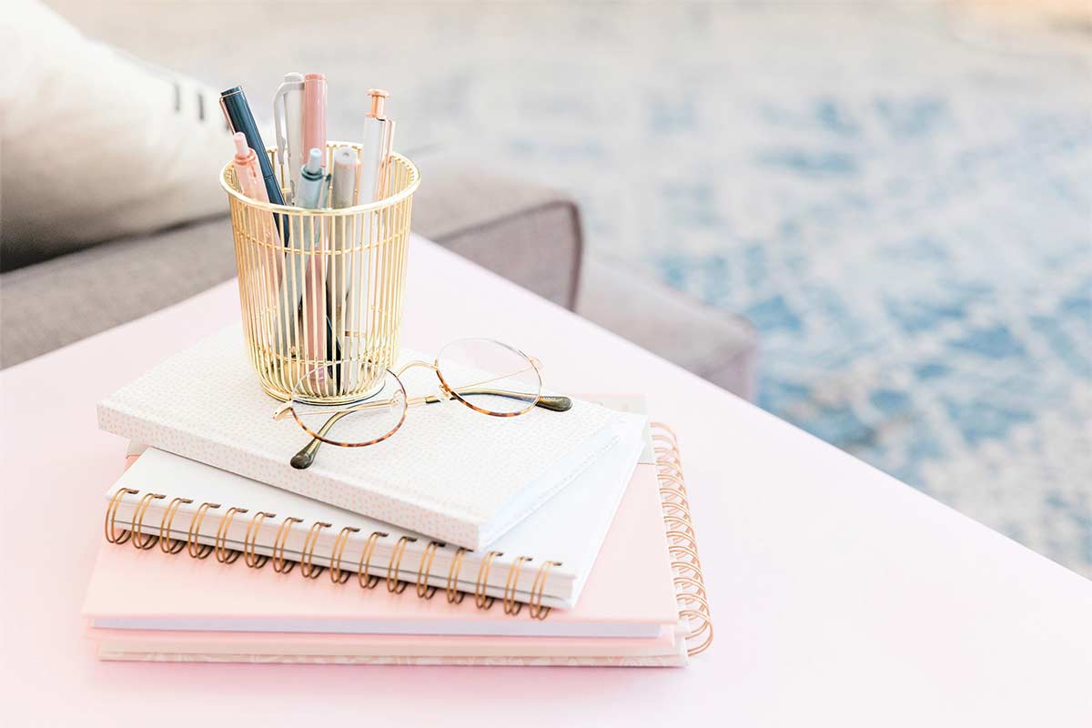 Pink-tinted table and journal stack with glasses and gold-tone pen holder.