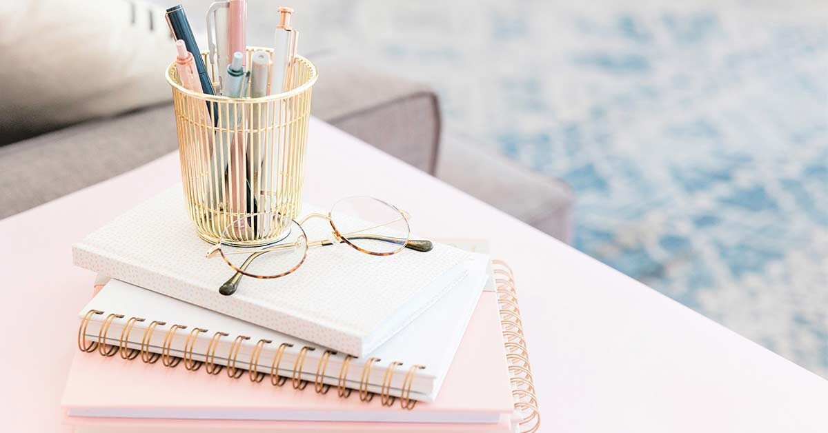 Pink-tinted table and journal stack with glasses and gold-tone pen holder.