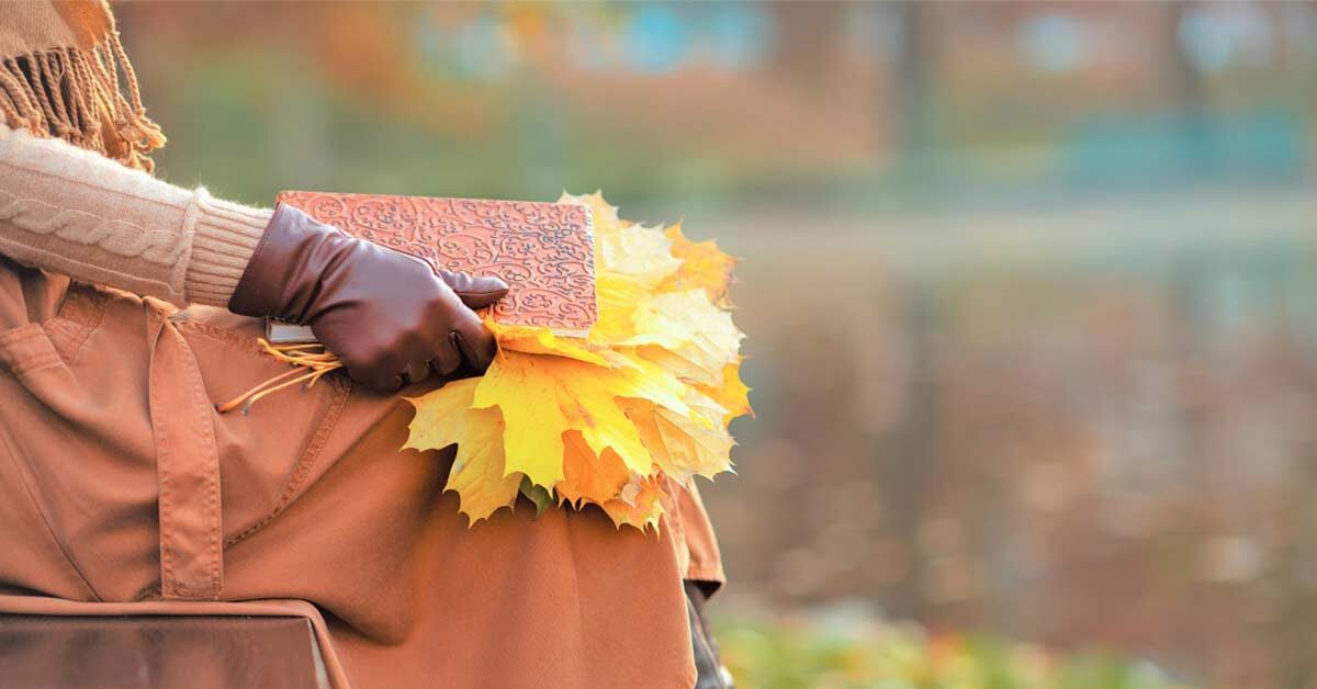 Woman in fall clothing and gloves sitting on bench and holding yellow leaves and notebook for writing fall journal prompts.