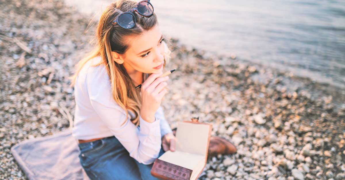 Woman with blond hair wearing blue shirt and jeans sitting on towel on stony beach while preparing to write responses to creative writing prompts for adults.