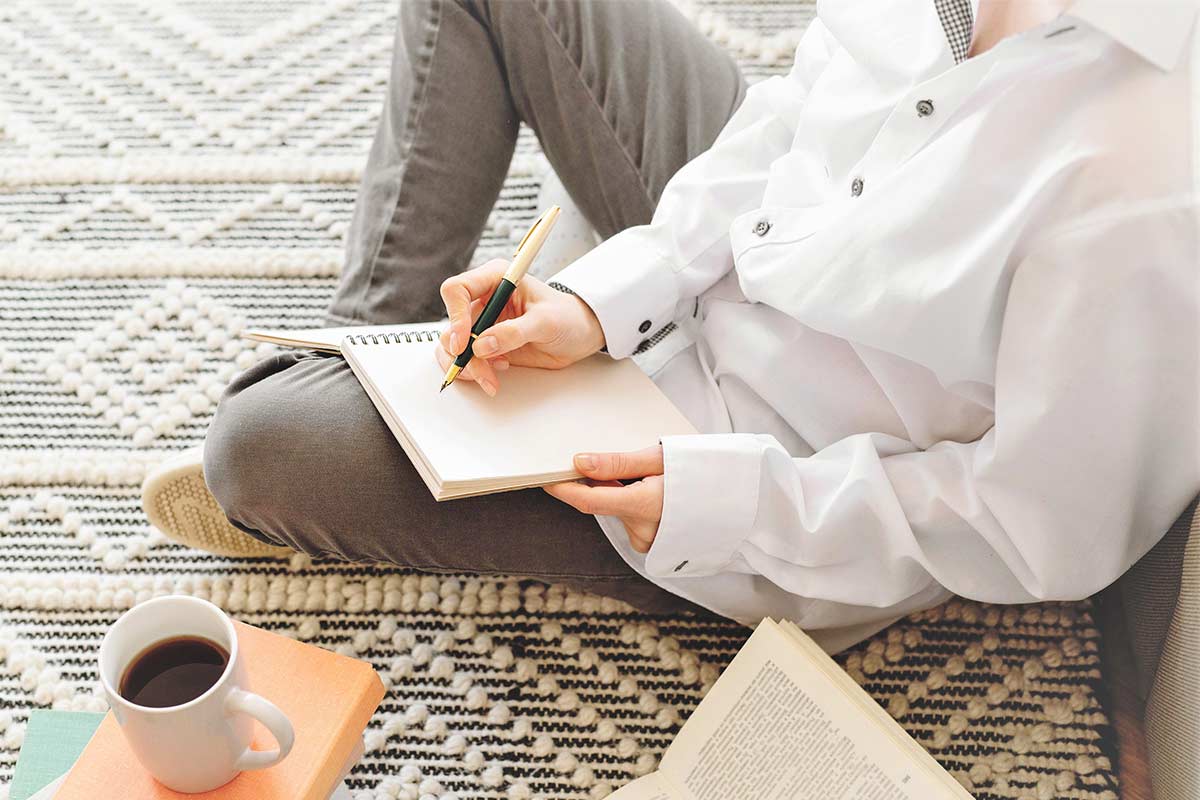 Woman in white shirt and black jeans sitting on floor with open notebook and preparing to use creative writing prompts.