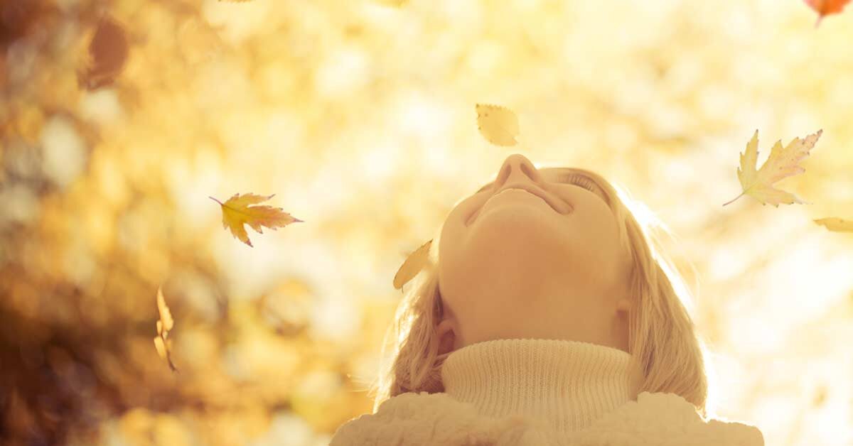 Blonde woman in turtleneck sweater smiling up at falling leaves on a sunny day.