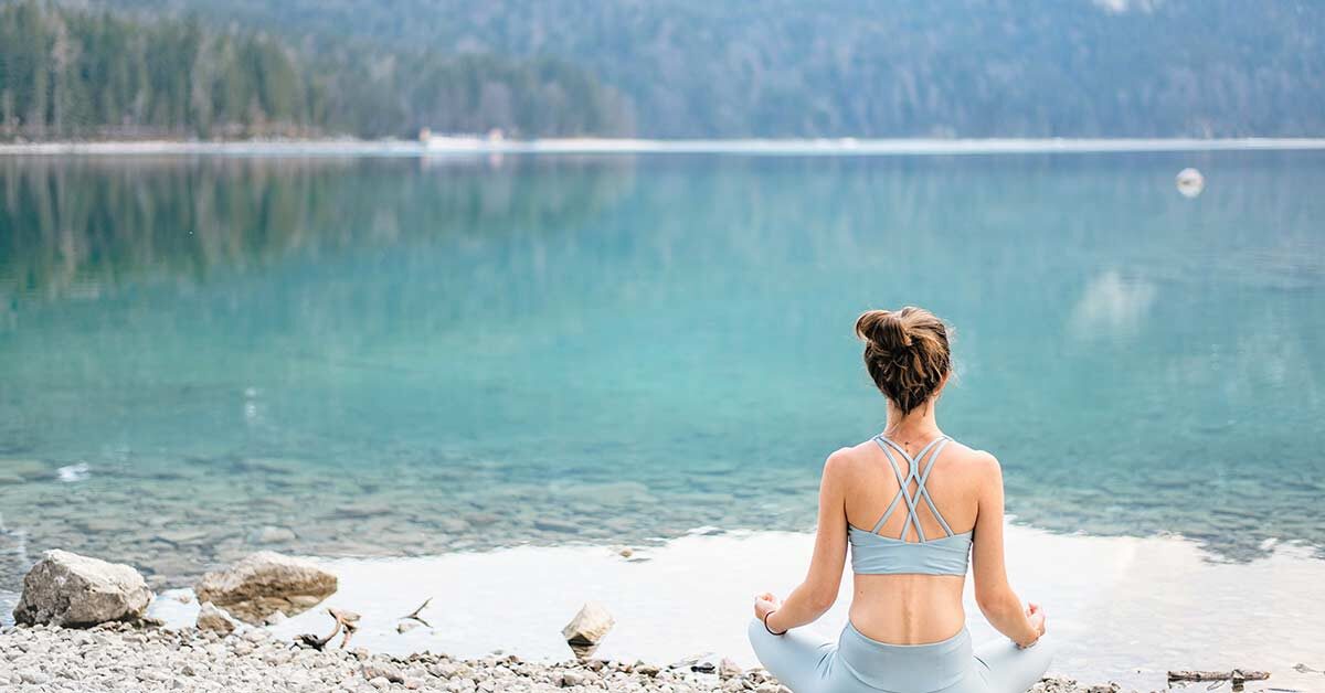 Woman wearing yoga outfit with hair pulled up sitting on stony beach taking time to slow down and breathe while looking at blue mountain lake.