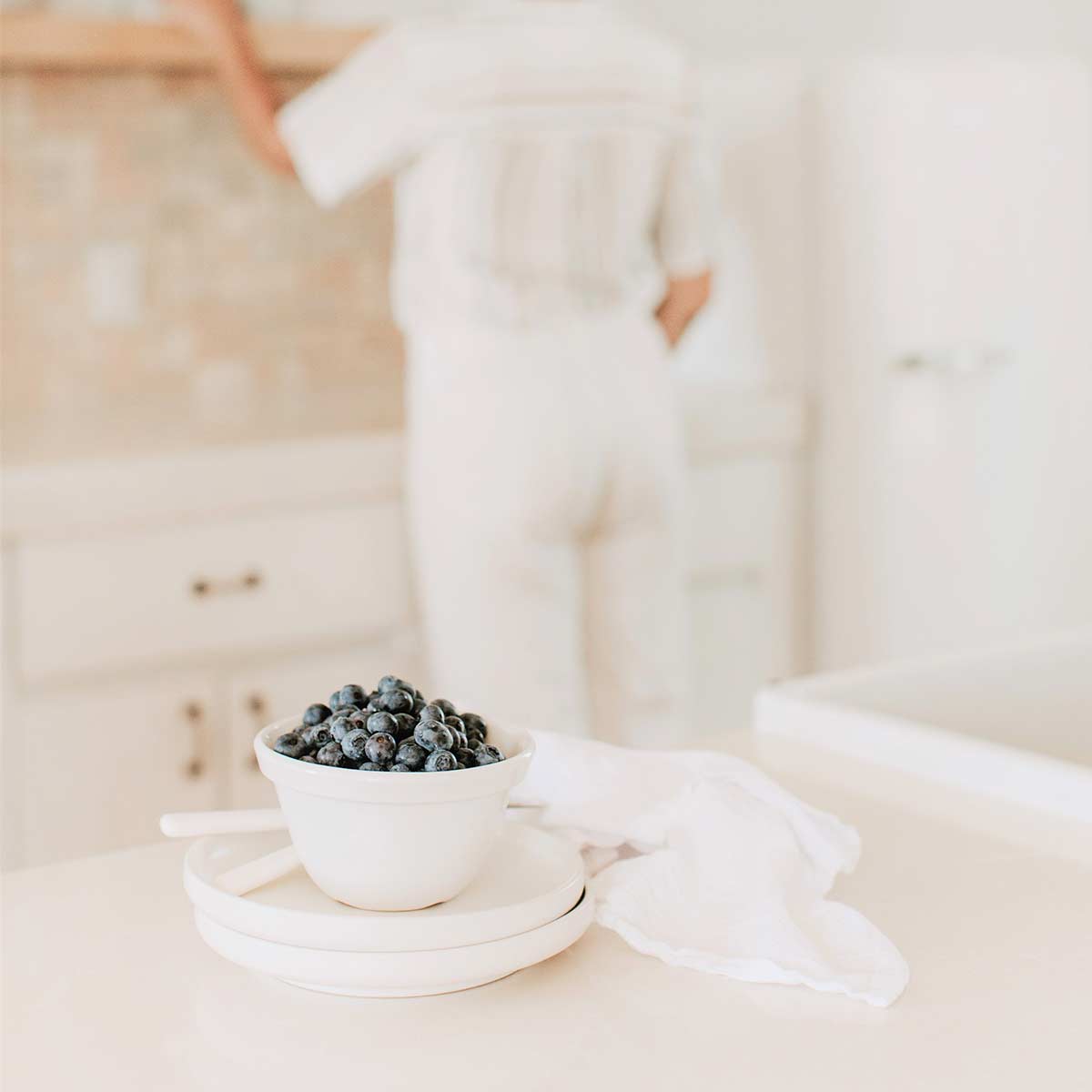 Bowl of blueberries on white dishes in white kitchen with blurred image of woman in background reaching for item on shelf.