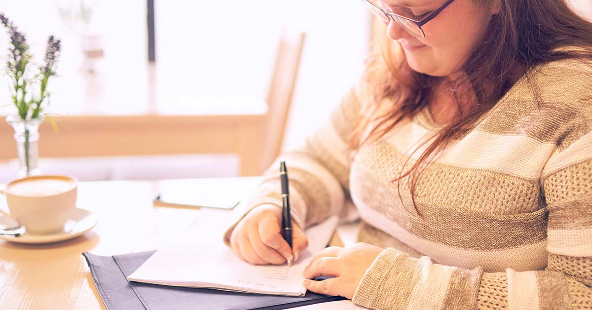 Woman with long dark hair wearing a sweater and glasses sitting at a table to do daily journal writing.