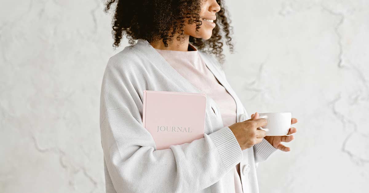 Woman in pink t-shirt and gray sweater holding pink journal and white coffee cup.