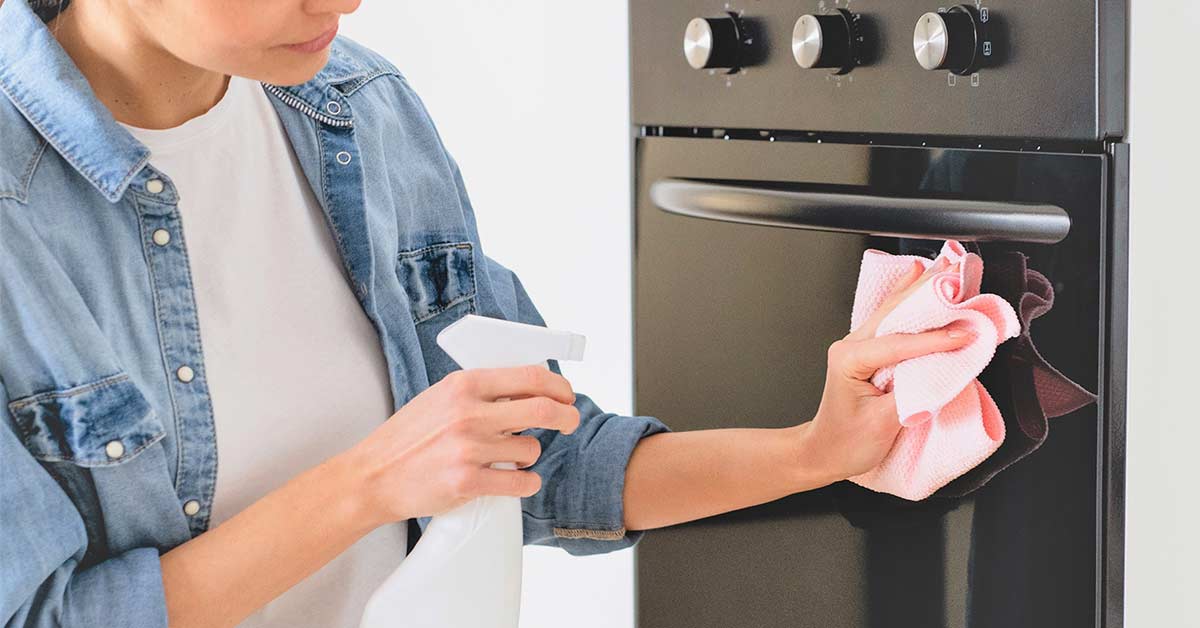 Woman wearing white t-shirt and denim shirt using spray cleaner to clean the front of her oven.