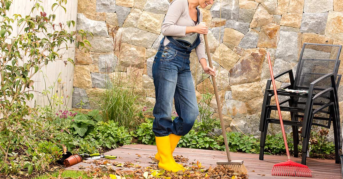 Woman wearing overalls and yellow boots sweeping deck as part of fall cleaning tasks.