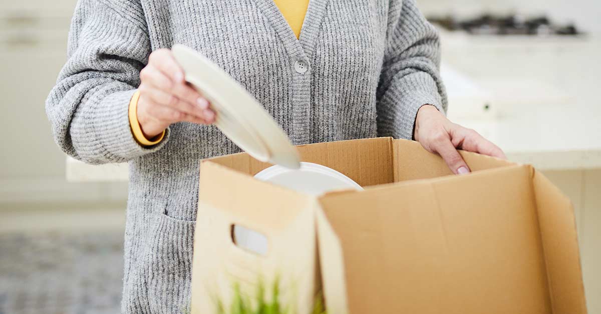 Woman wearing gray sweater and sorting dishes into cardboard box.