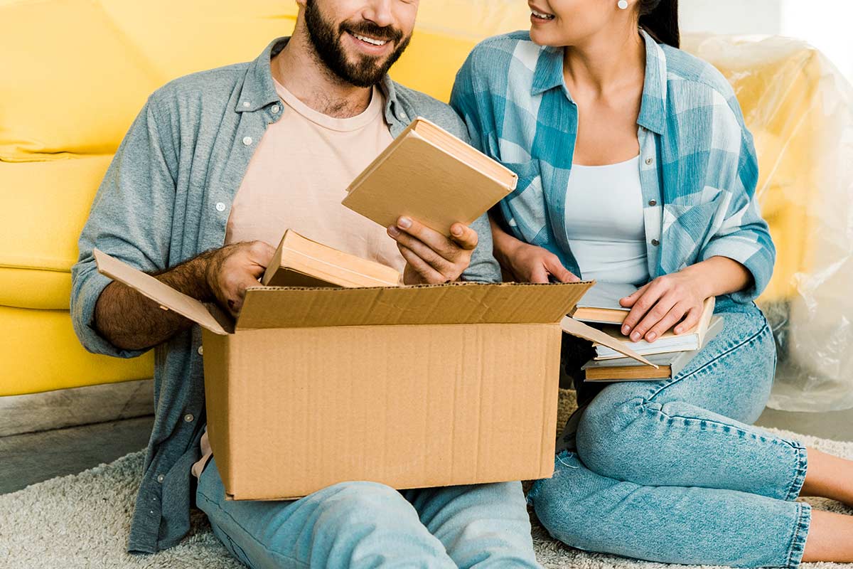 Man and woman wearing casual shirts and jeans sitting on floor in front of yellow couch and sorting books into cardboard box.