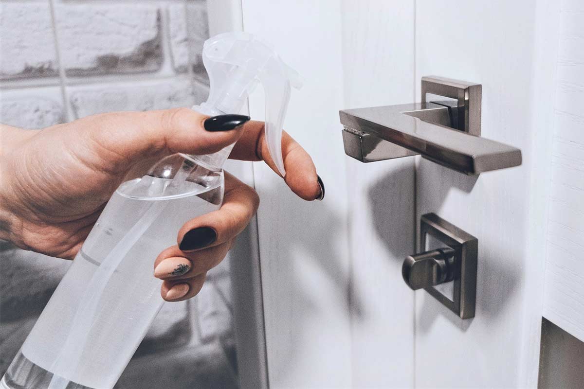 Woman's hand holding spray bottle to clean door handle on white entry door.