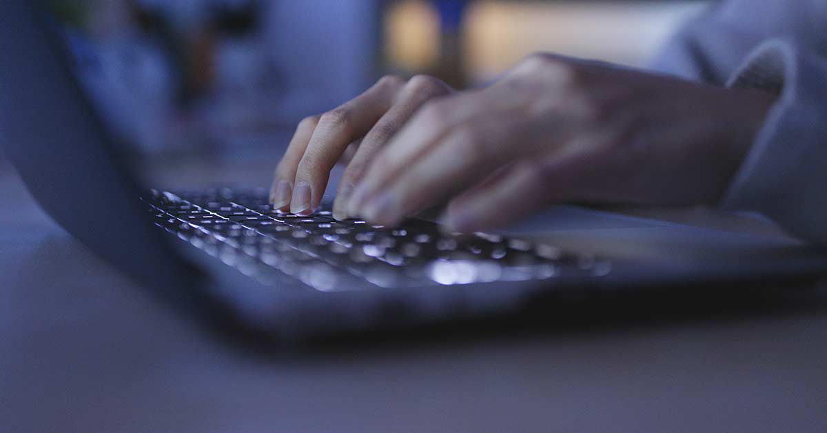 Female hands typing on backlit laptop keyboard in dark room.