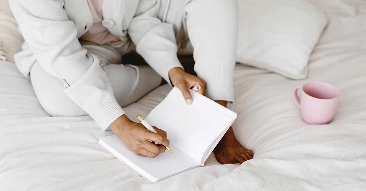Woman wearing soft gray clothes sitting on bed with pink coffee mug preparing to write in journal.