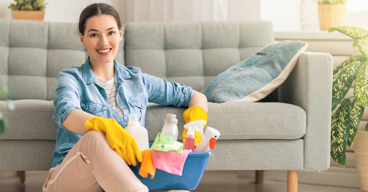 Woman with dark hair and chambray shirt sitting on floor in front of gray couch while resting from cleaning the house and holding basket of cleaning supplies.