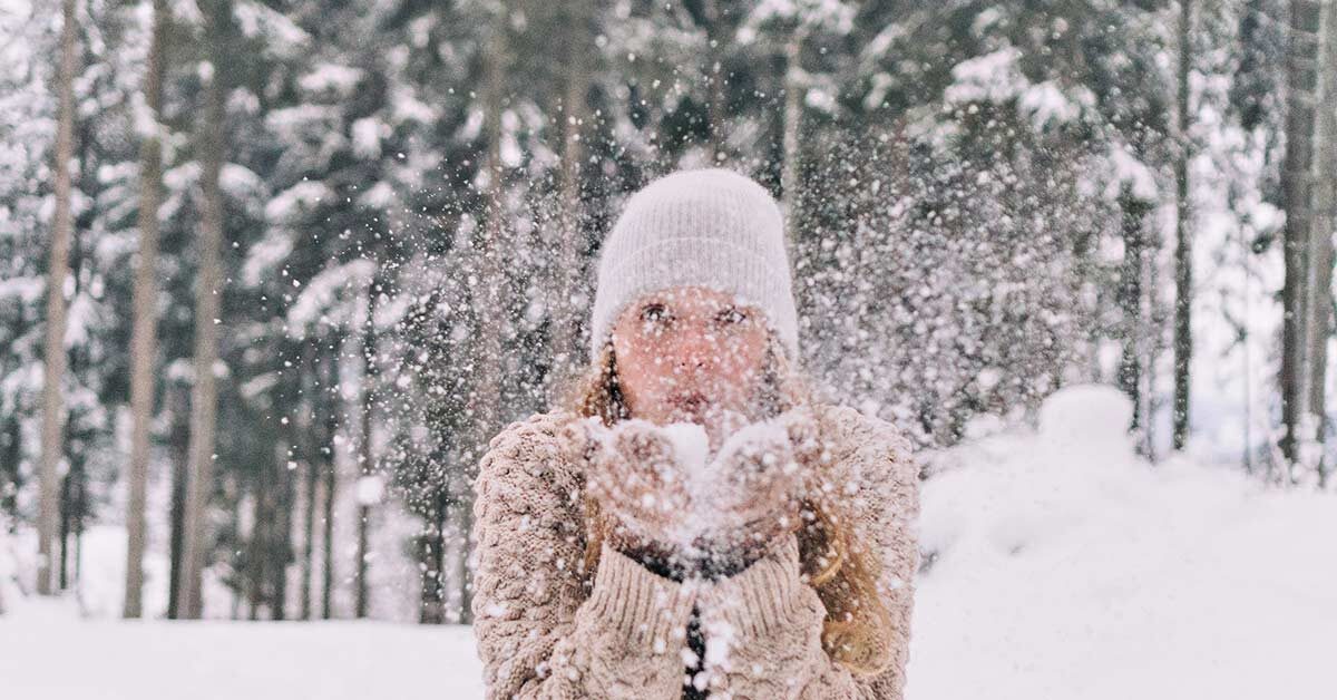 Outdoor snowy background with woman in sweater, mittens, and hat blowing snow.