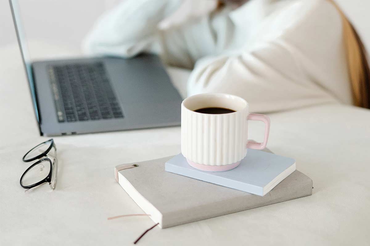 Woman in white sweater preparing for early morning writing while sitting at white table with laptop, journals, coffee and glasses.