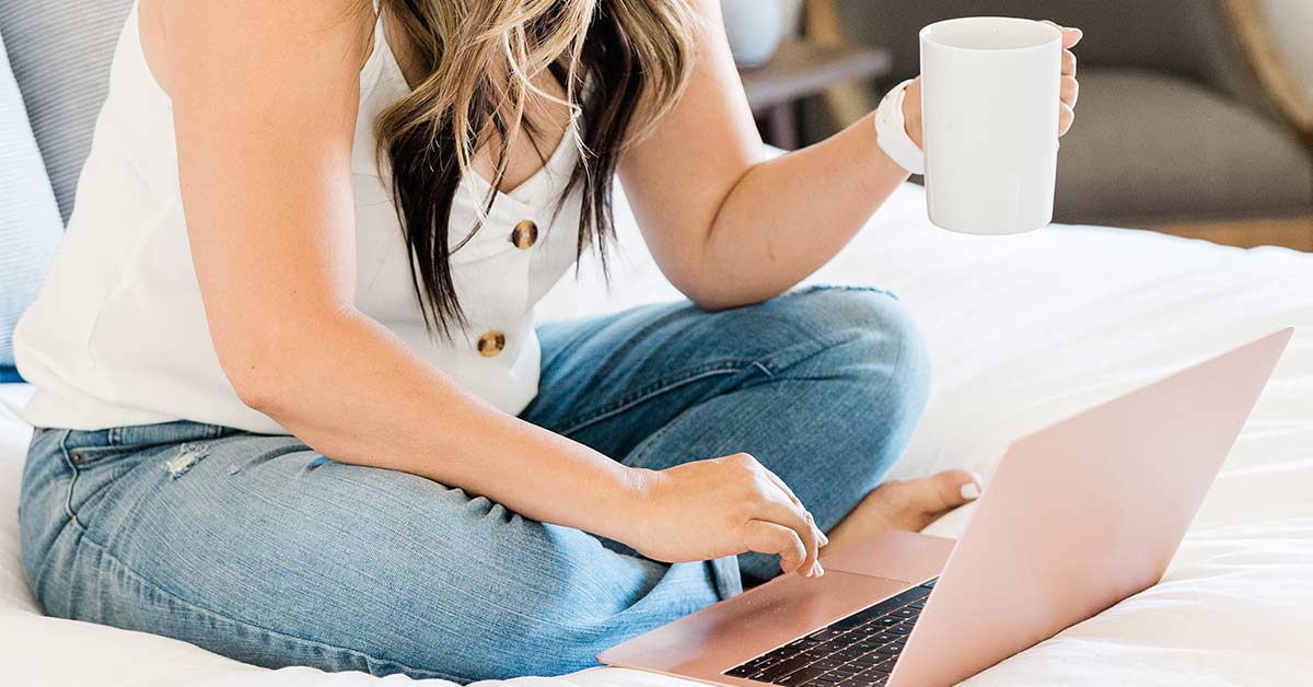 Woman wearing white tank top and jeans sitting on bed and holding coffee mug while typing on laptop computer.