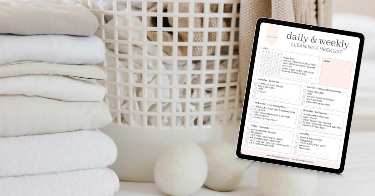 Image of daily and weekly home cleaning schedule printable over background of laundry basket next to dryer balls and pile of folded clothes.