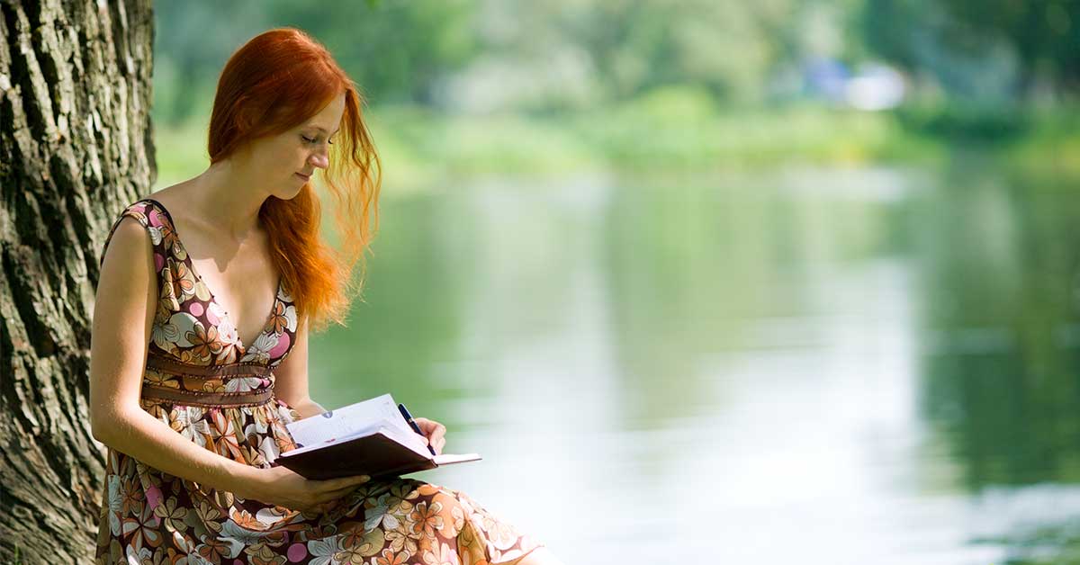 Red-haired woman writing in journal while sitting outdoors.