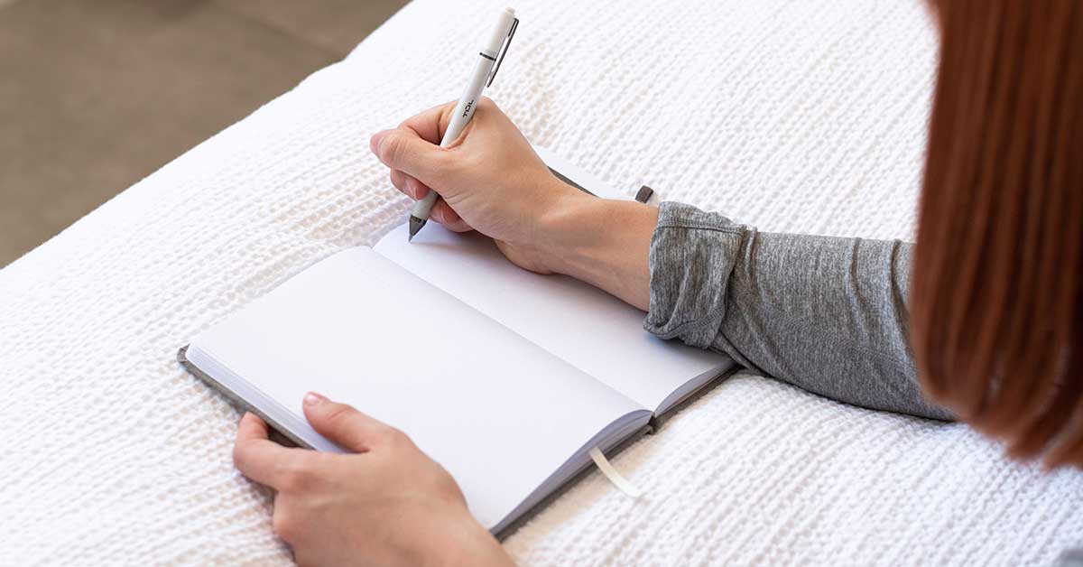 Woman with red hair preparing to do creative writing in journal while lying on white blanket.