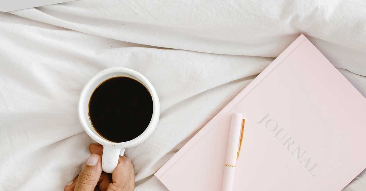 Pink journal and pen next to female hand holding cup of coffee on white background.