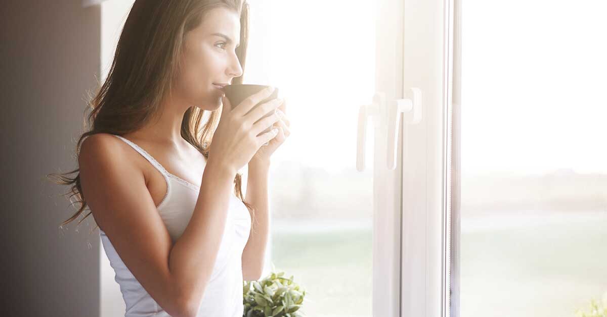 Woman standing at sunny window and sipping a beverage.