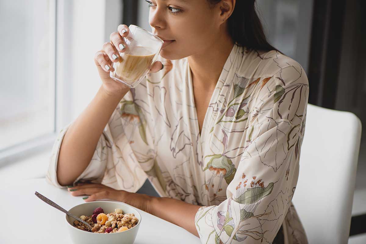 Young black woman wearing a botanical-pattern robe and drinking from a glass while sitting at a breakfast in a sunny room.