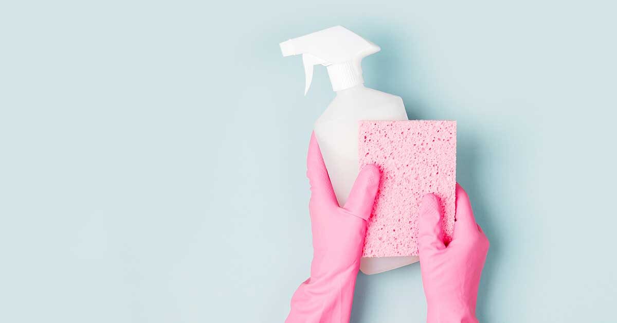 Hands in pink gloves holding pink cleaning sponge and white spray bottle against light blue background.