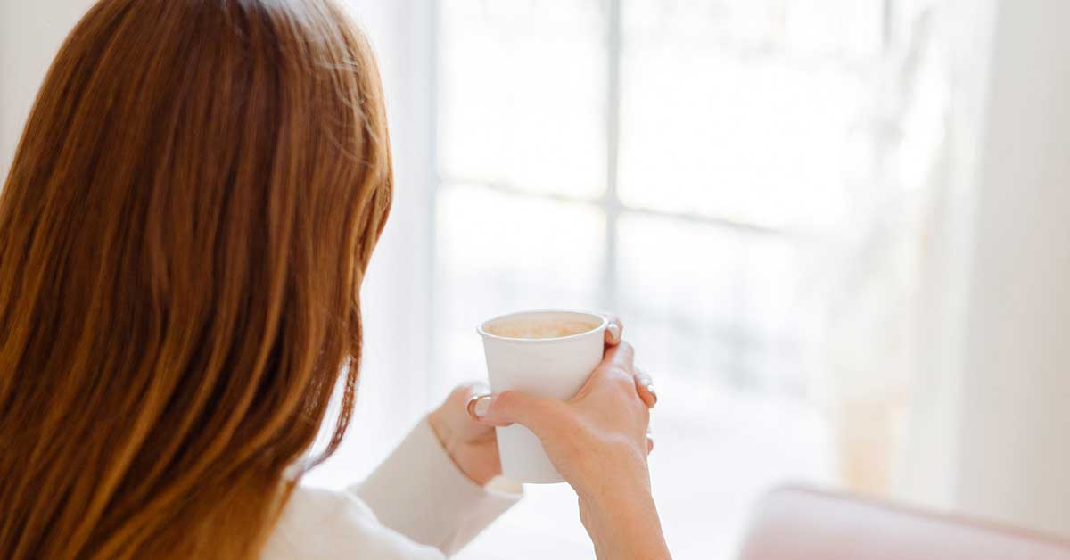 Red-haired woman holding mug and gazing away from camera toward window.