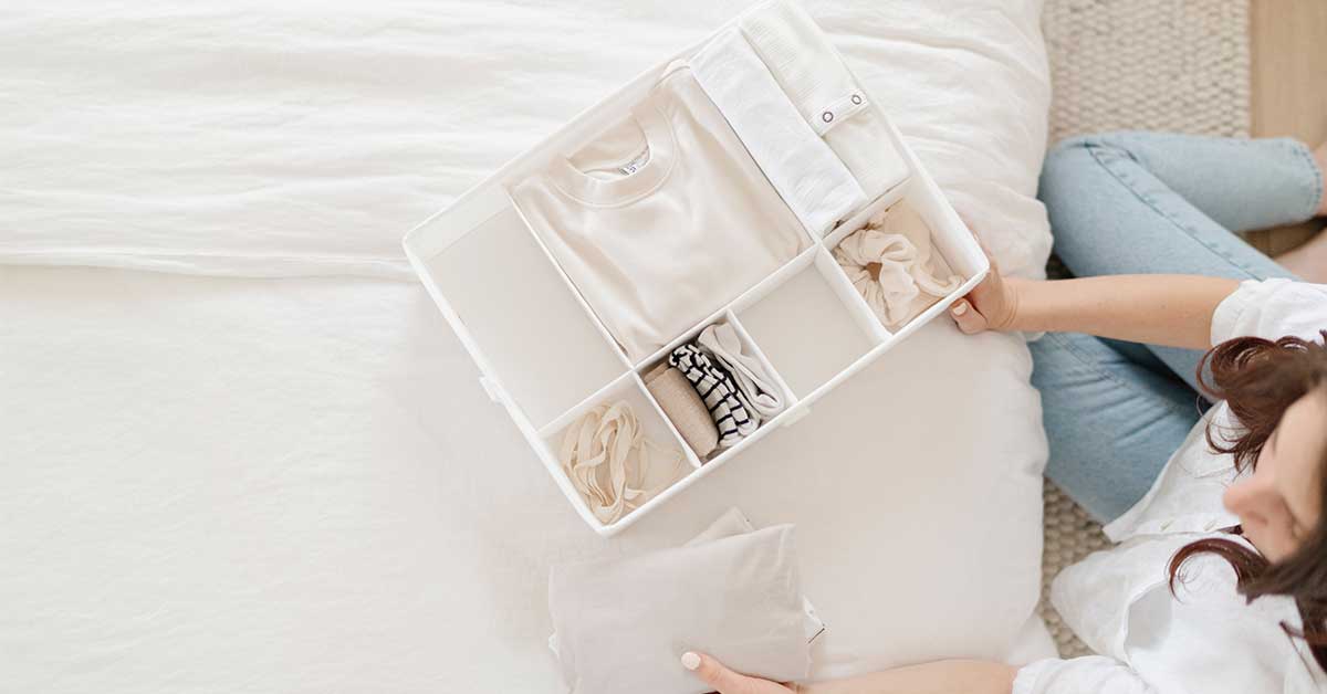 Woman sitting on floor next to bed while organizing clothes in a storage container.