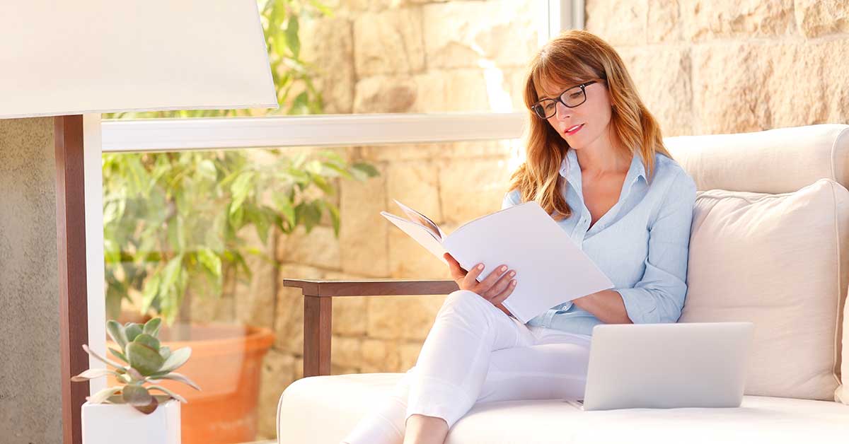 Midlife woman sitting on white sofa browsing booklet with white cover.