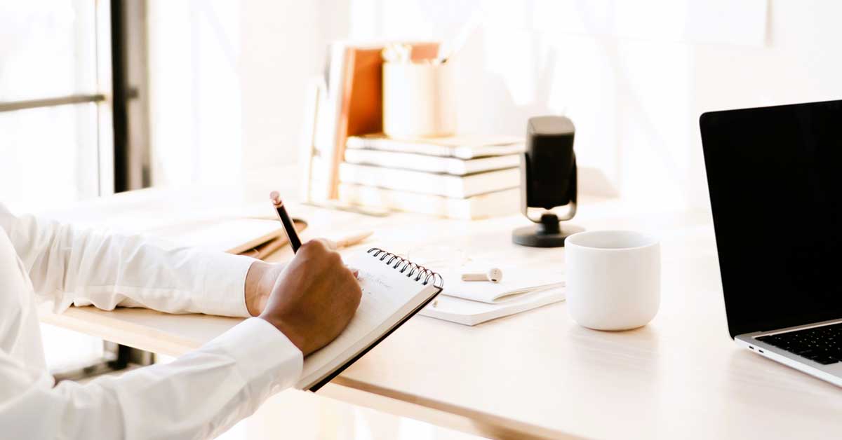 Hands holding notebook and pencil at white desk in sunny room.