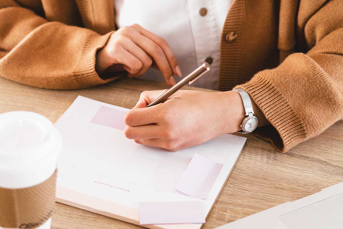 Partial image of woman sitting at desk and writing on a planner notepad with sticky notes.