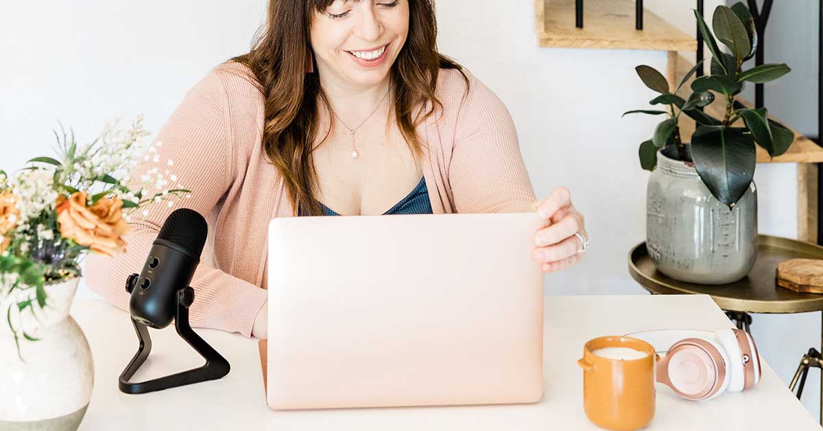 Woman sitting at desk with open laptop next to microphone in front of white and wood staircase.