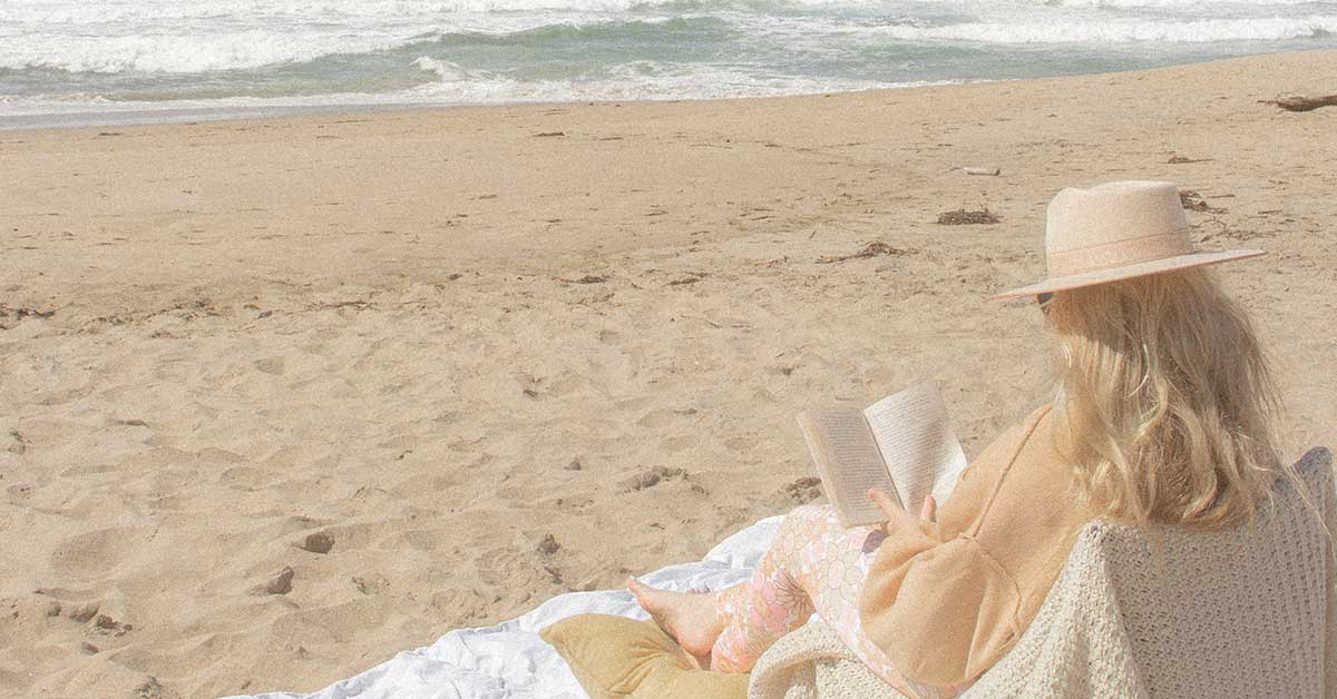 Woman with blond hair and hat sitting on blanket and reading at the beach.