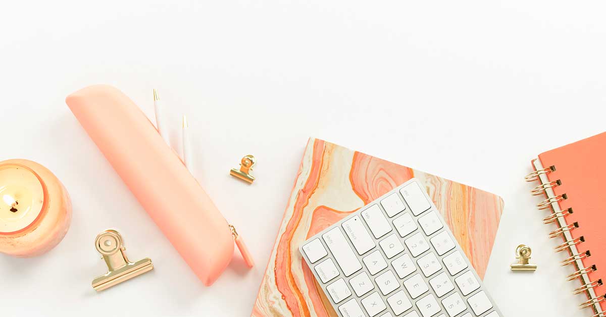 Overhead shot of coil-bound planner, notebook, keyboard, pencil case, candle and clips arranged on white desk.