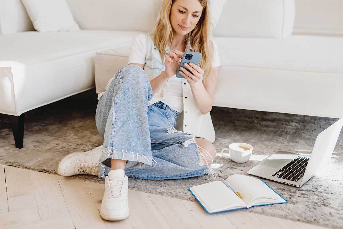 Woman with blond hair sitting on living room floor and working on phone next to open laptop and notebook.