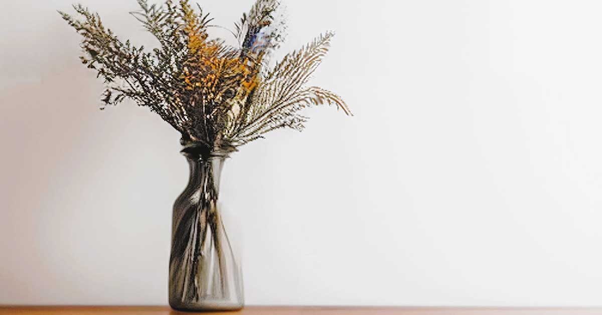 Glass vase of decorative dried plants on clutter-free wood shelf against white wall.