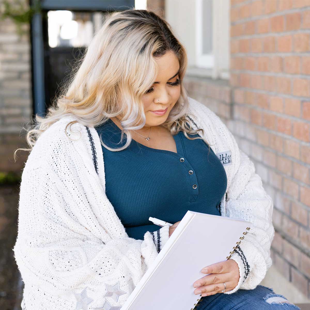 Woman with blond hair wearing white sweater and blue shirt sitting outside and writing in journal.