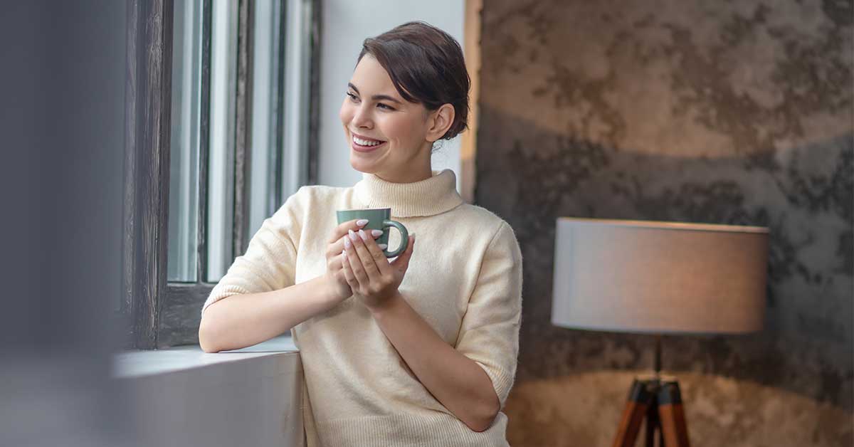 Smiling woman holding mug while gazing out window.