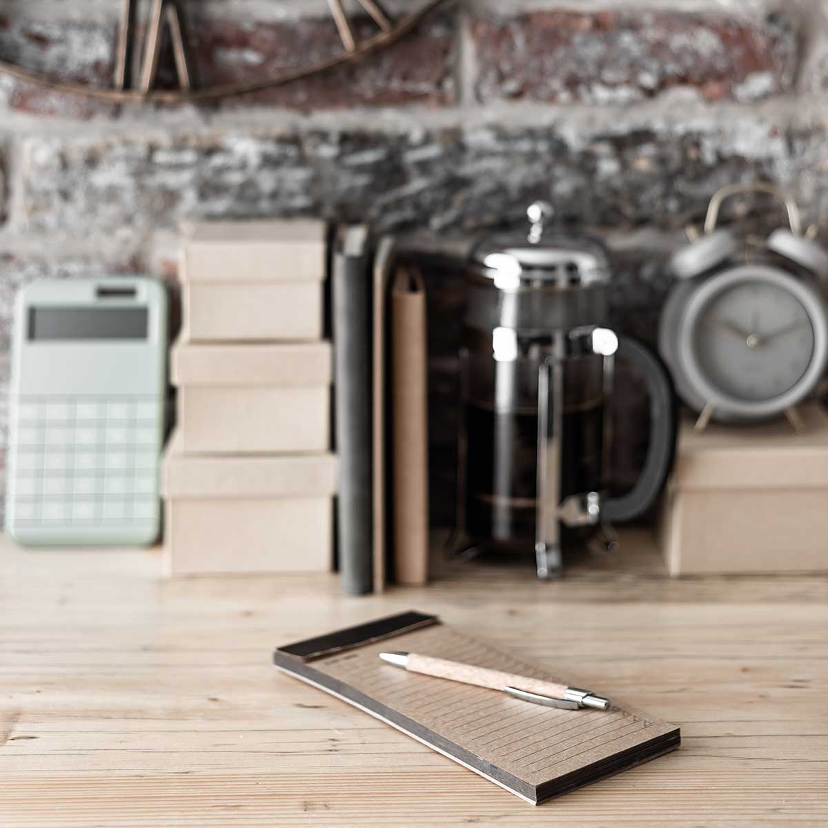 Notepad, boxes, and coffee pot on wood counter in front of brick wall.