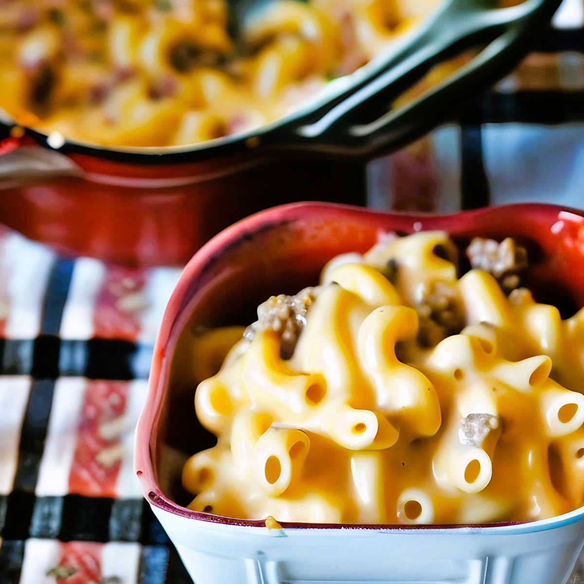 Macaroni cheeseburger casserole serving in ceramic dish with red rim and blurry background of plaid tablecloth and main casserole dish.