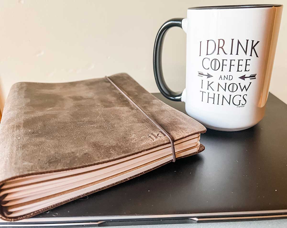 Leather journal and coffee mug sitting on closed laptop computer in front of pale yellow wall.